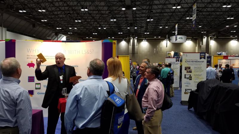 Magic Scott building a crowd at the Library Systems & Services booth at the ICMA conference, 2016 — six attendees gathered around the booth watching a sleight-of-hand routine