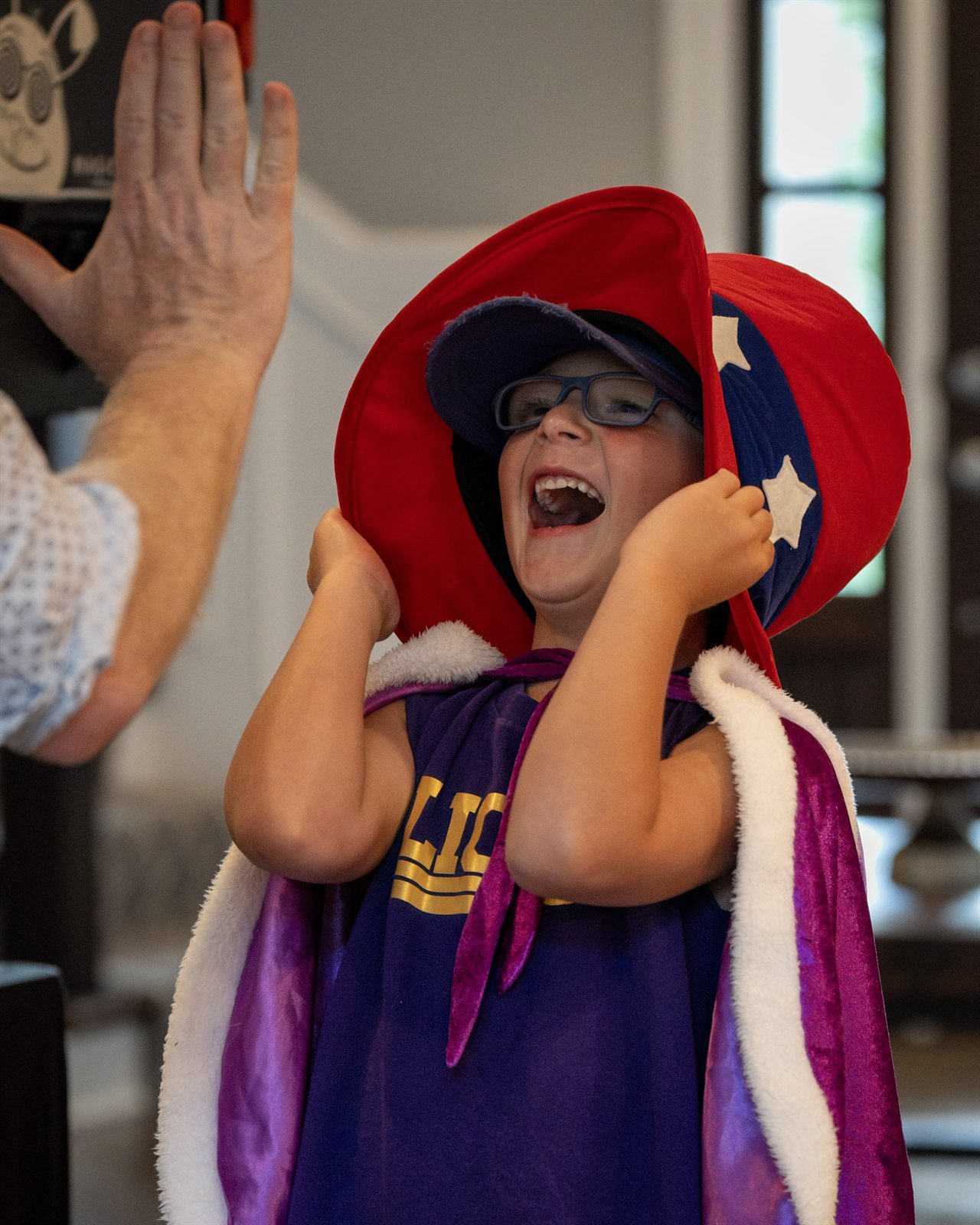 Birthday-party kid in an oversized magician's cape and star-spangled wizard hat, mid-laugh, high-fiving Magic Scott at a Kansas City home party