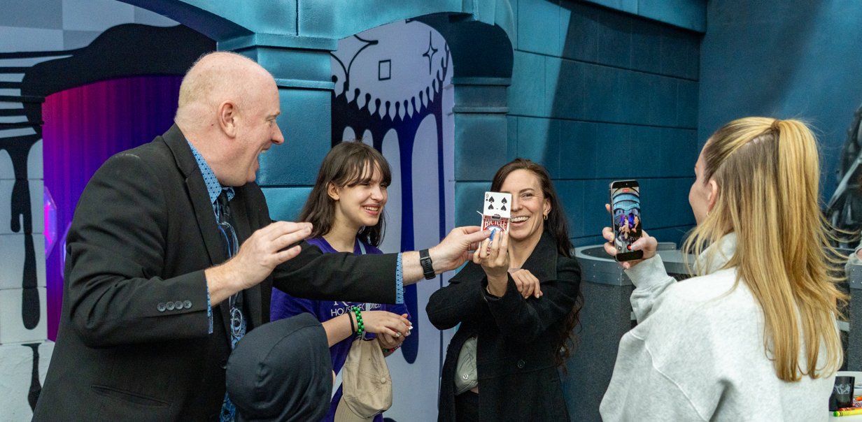 Three young adults at a Science City after-hours event, holding playing cards up to their faces with delighted reactions, while Magic Scott looks on