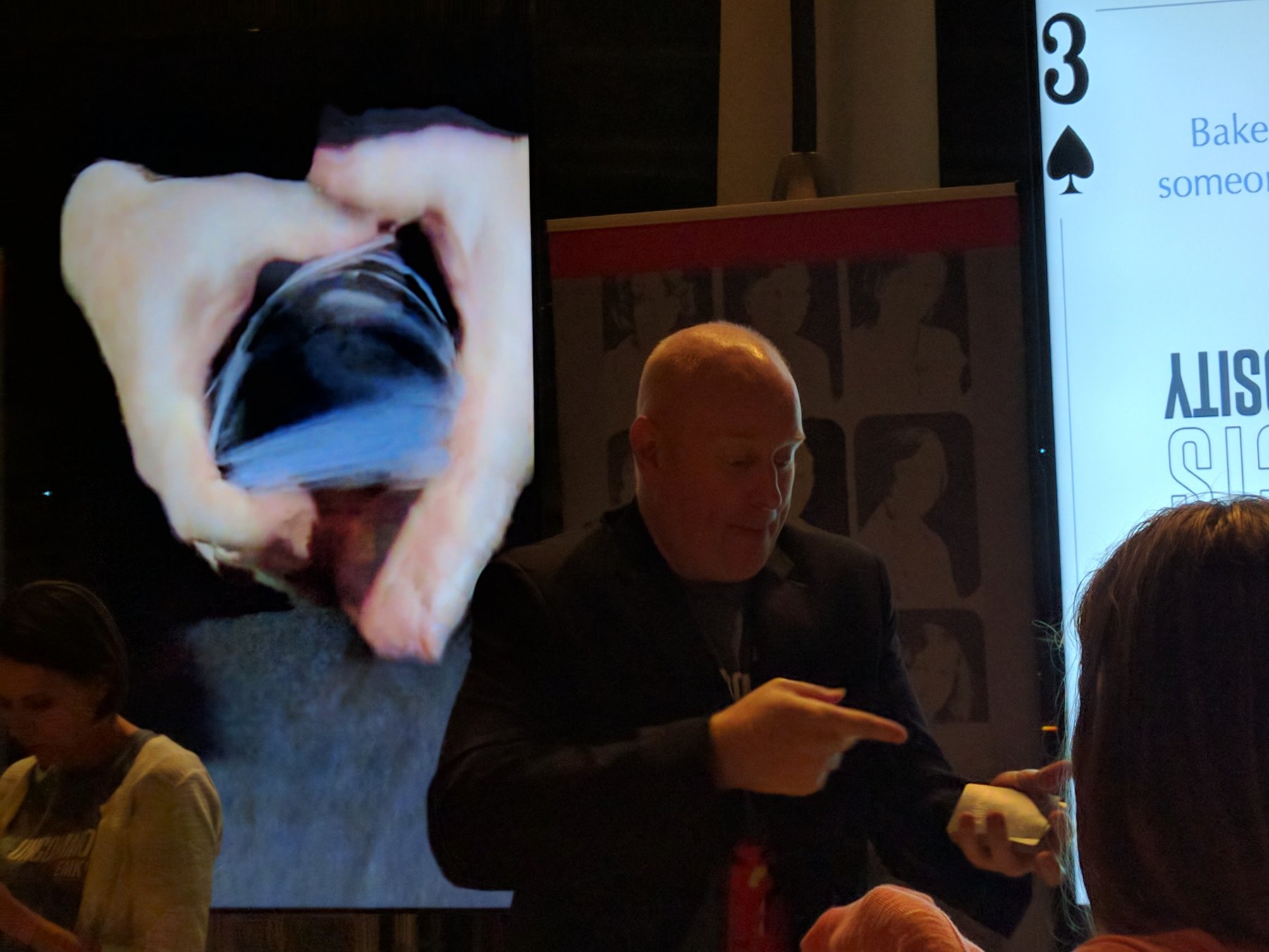 Magic Scott on stage with a live close-up camera projecting his hands onto a giant screen behind him — a card trick magnified for the back row
