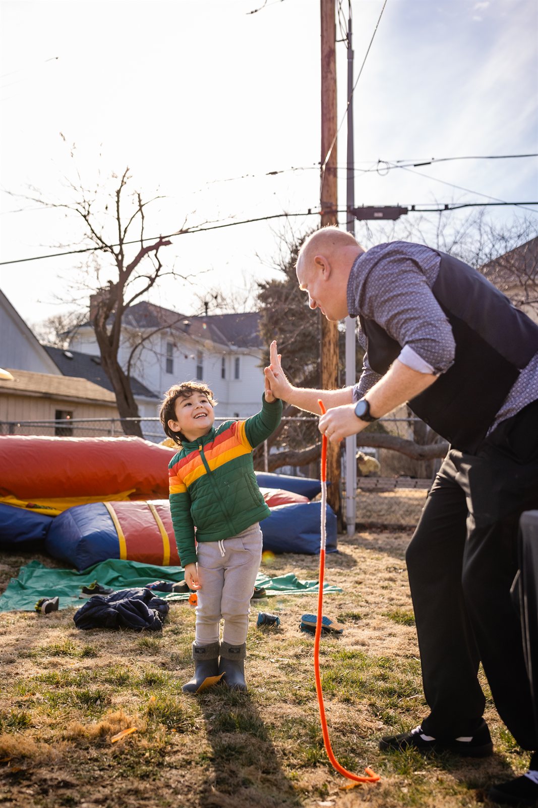 Magic Scott bowing toward a small boy in a striped sweater for a high-five at an outdoor backyard birthday, bounce house behind.
