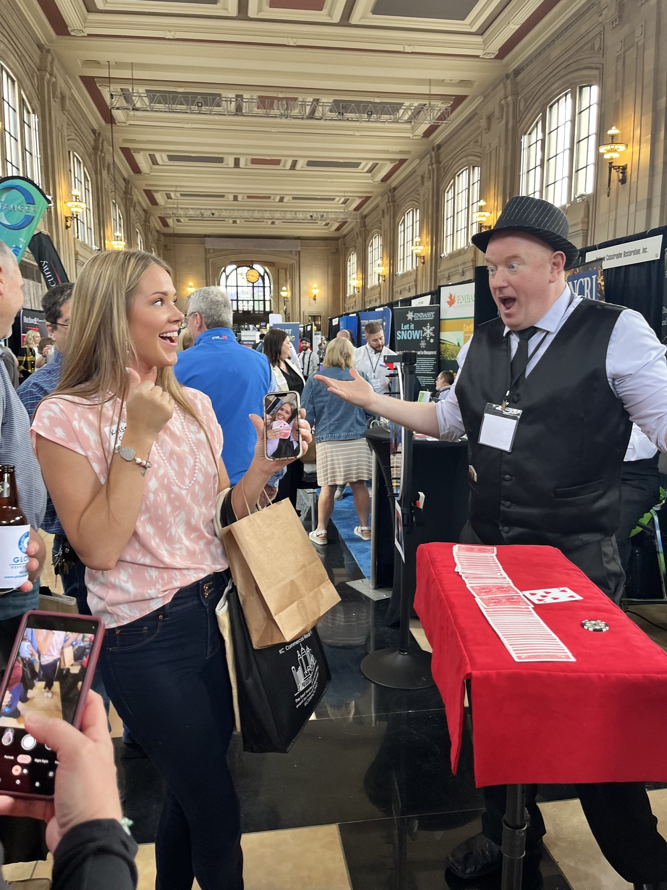 Magic Scott in a derby cap and vest engaging two booth visitors at a Union Station Kansas City trade show — a woman in pink mid-laugh while a friend captures the moment on a phone