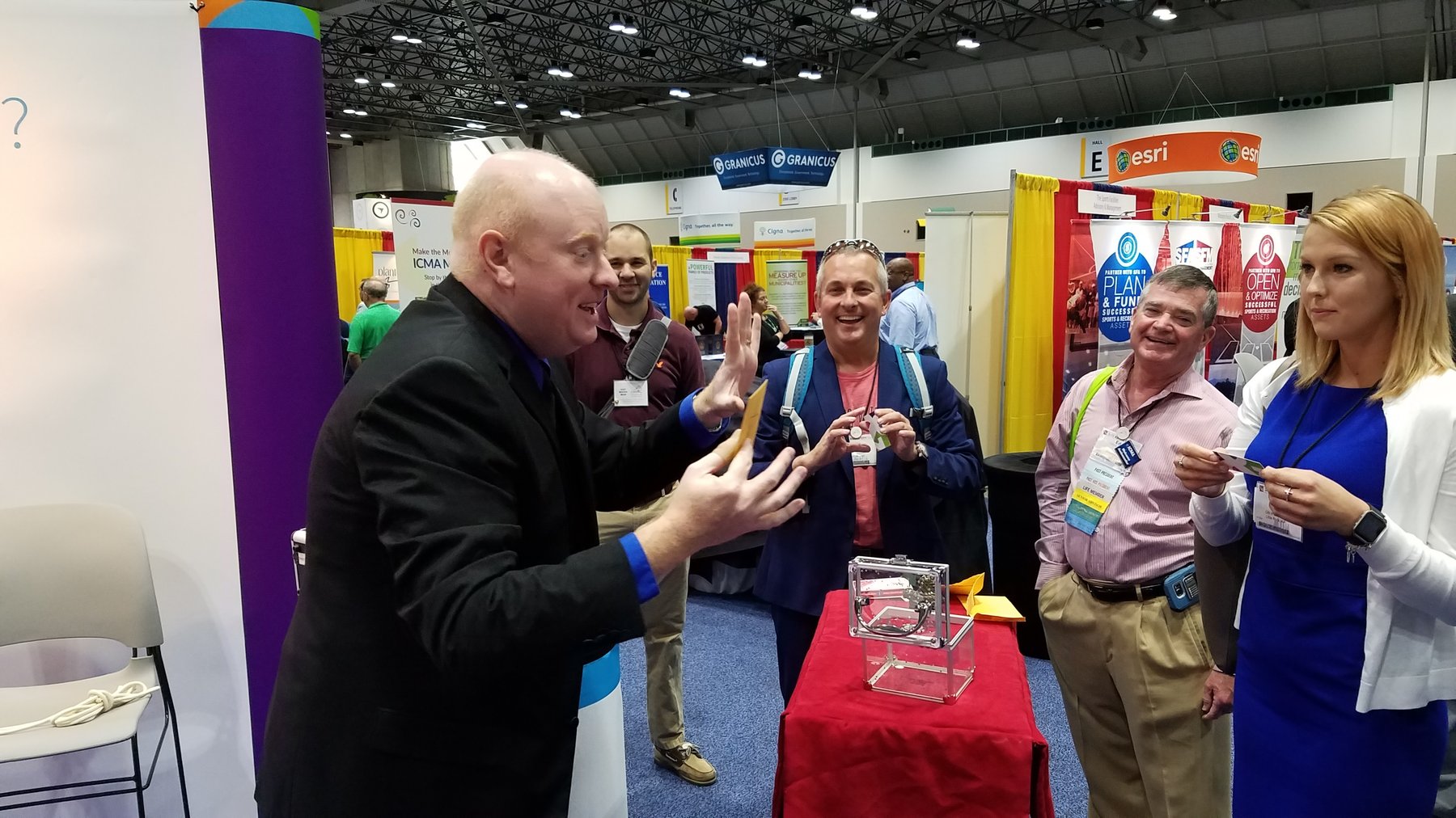 Magic Scott mid-routine at the Library Systems & Services booth at the ICMA conference, 2016 — three booth visitors watching a close-up effect with delighted reactions