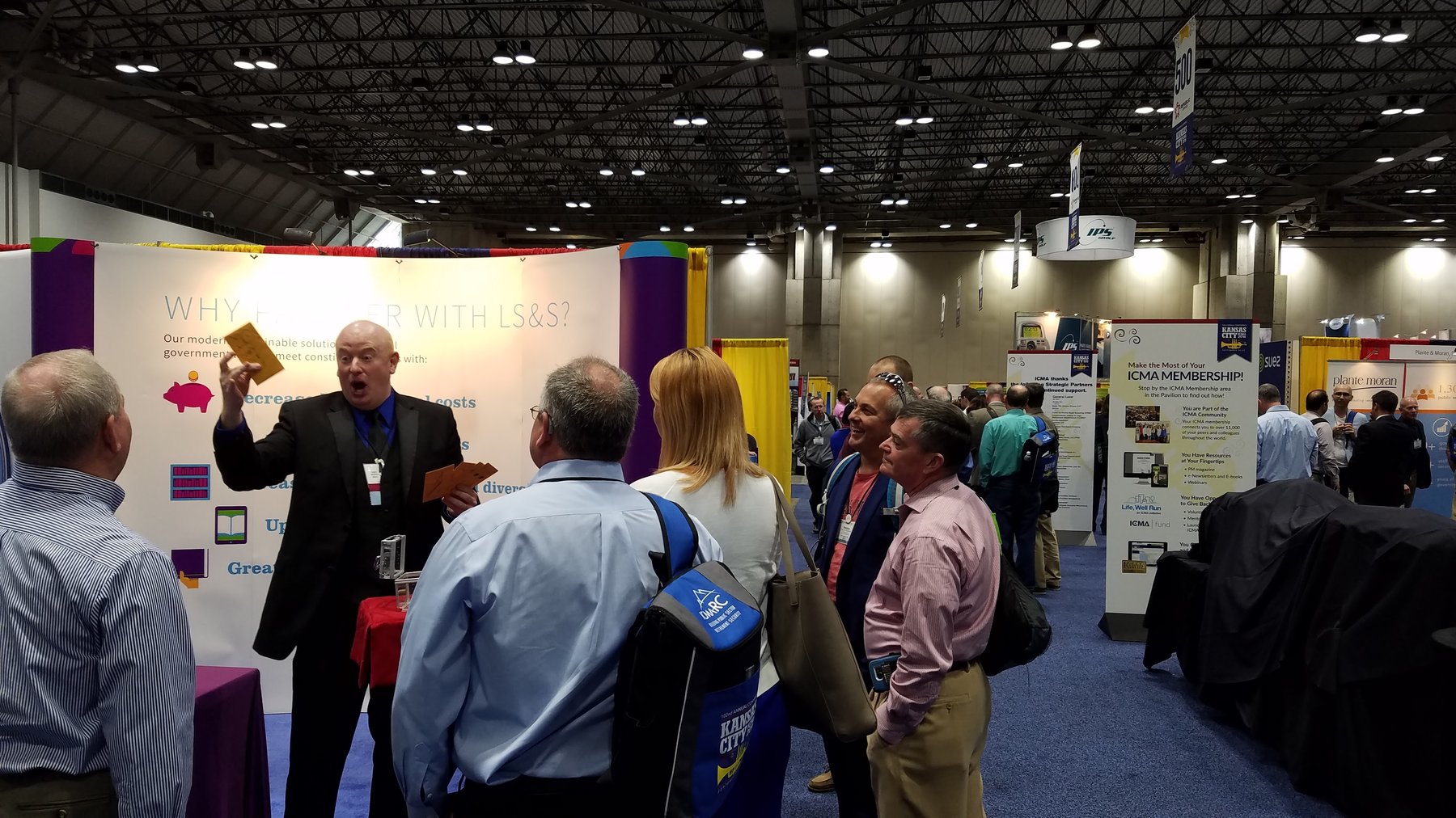 Magic Scott building a crowd at the Library Systems & Services booth at the ICMA conference, 2016 — six attendees gathered around the booth watching a sleight-of-hand routine